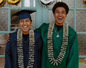 two happy young men in graduation robes and wearing paper money leis